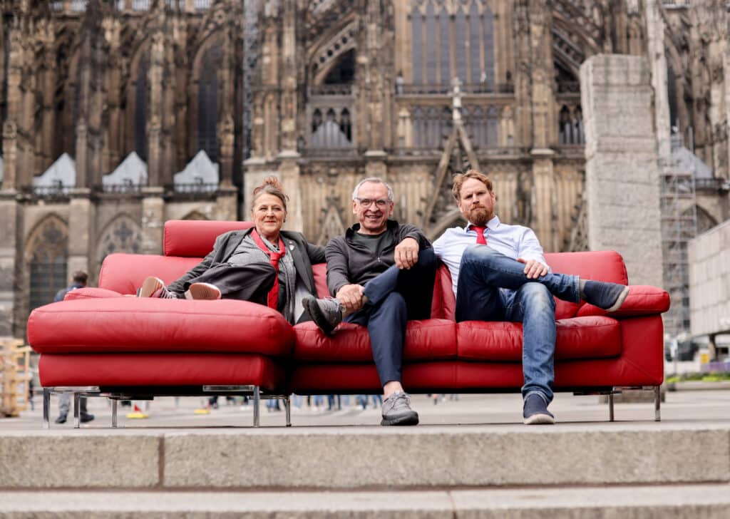 Die FRAKTION im Rat der Stadt Köln: Die Ratsmitglieder Birgit Beate Dickas, Walter Wortmann und Michael Hock (v.l.n.r.) posieren auf einer roten Couch vor dem Weltkulturerbe Kölner Dom